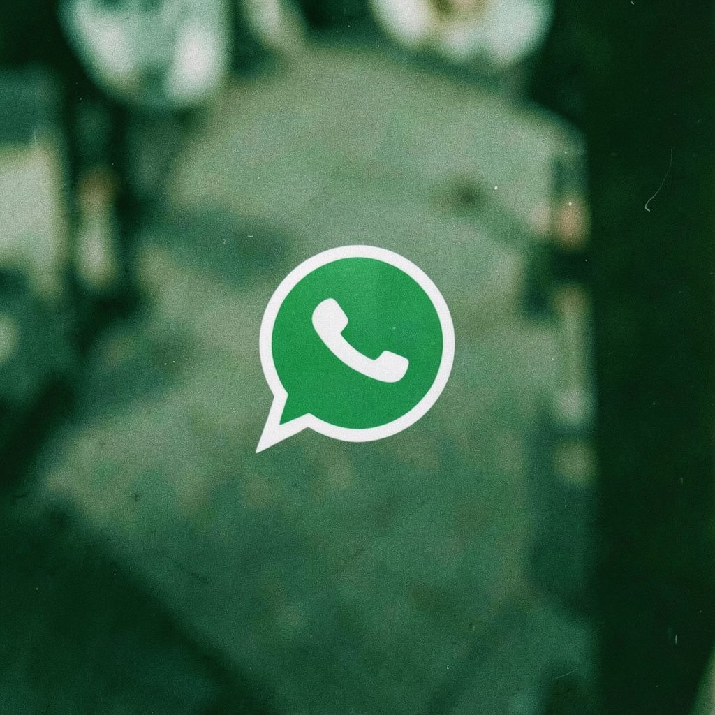 Smartphone screen showing a WhatsApp chat interface with a green header and message bubbles, held in a hand against a neutral desk background.